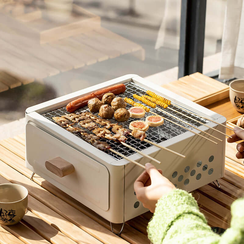 White square barbecue oven outdoors with grilled food, suitable for 3-5 people, on wooden table.